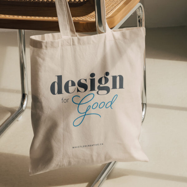 A canvas tote bag with the text "design for good" imprinted on it, resting against a metal chair in sunlight.
