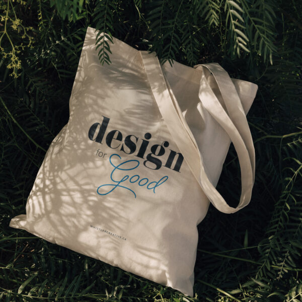 A Design for Good sustainable tote bag, resting among green leaves.