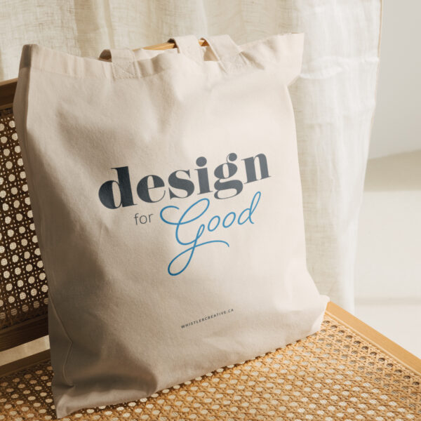 Design for Good sustainable tote bag with "design for good" text rests against a wooden chair with a sheer curtain backdrop.
